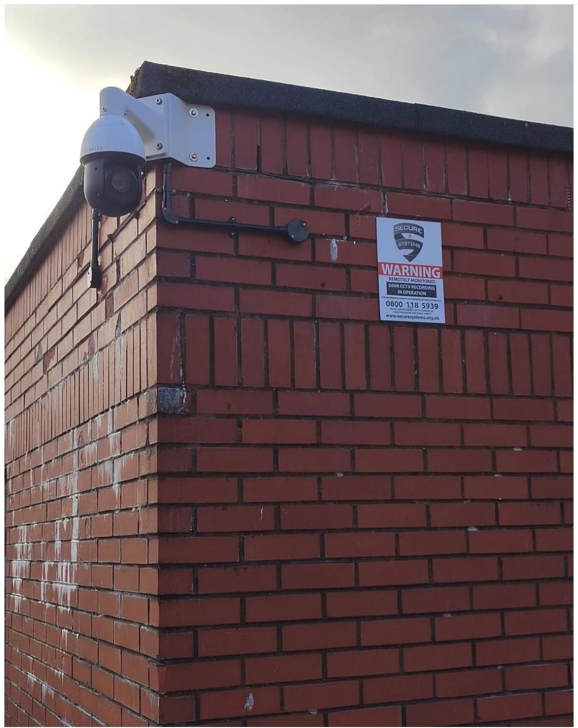 A dome camera and a CCTV warning sign fitted high at the corner of a brick building.