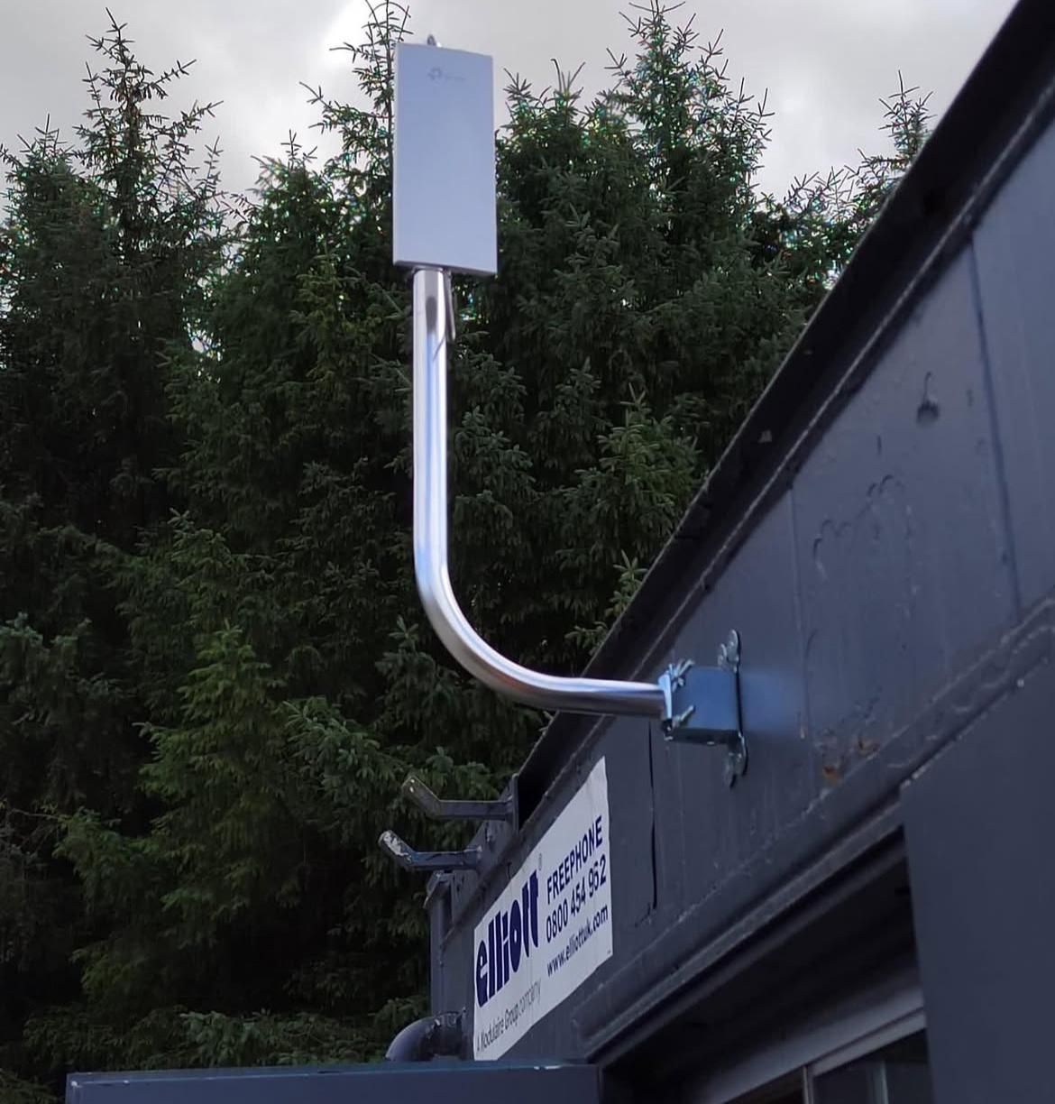 A point-to-point wireless bridge on a bracket, aimed across a stand of conifers.